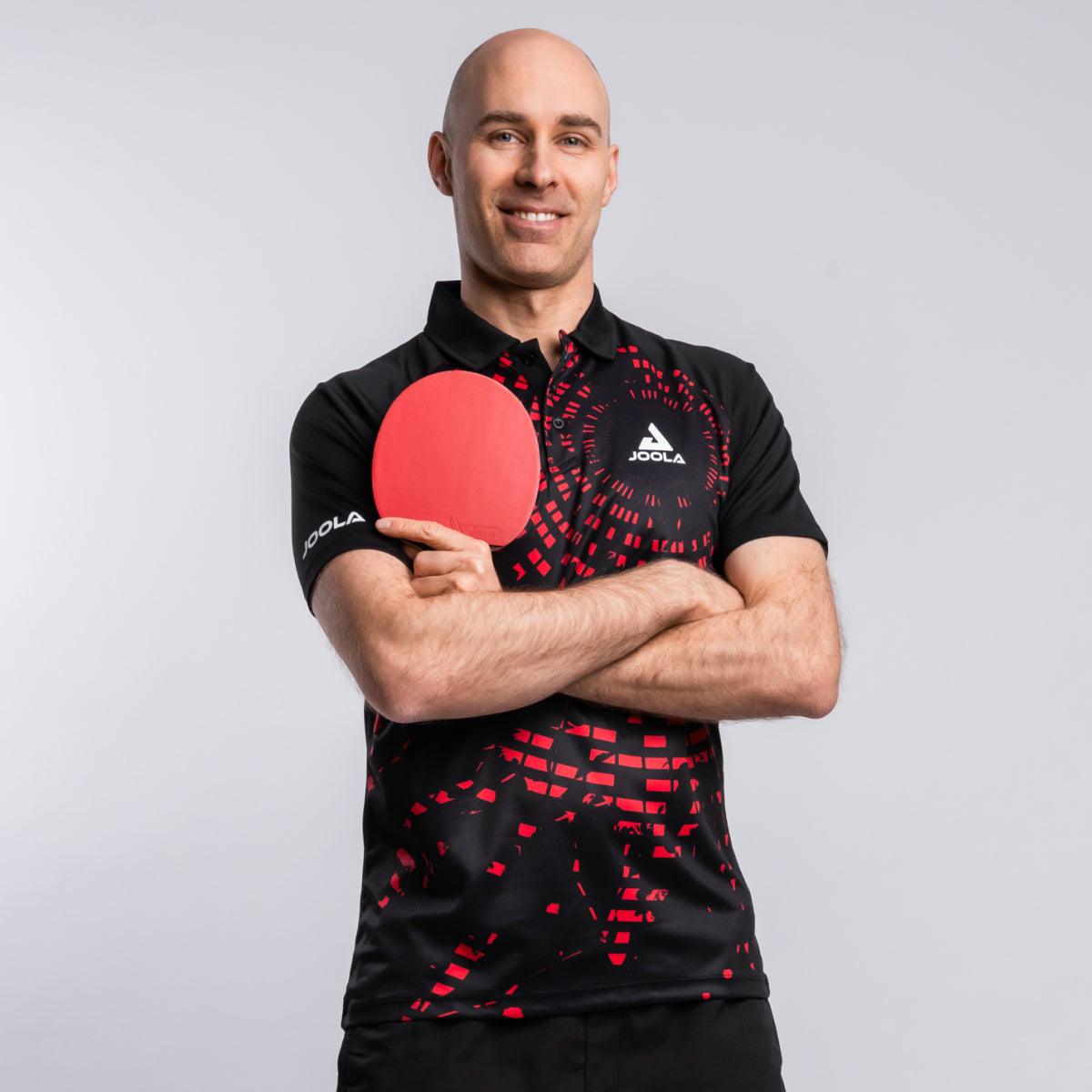 Mann in schwarzem JOOLA-Poloshirt mit rotem Muster hält roten Tischtennisschläger vor weißem Hintergrund.