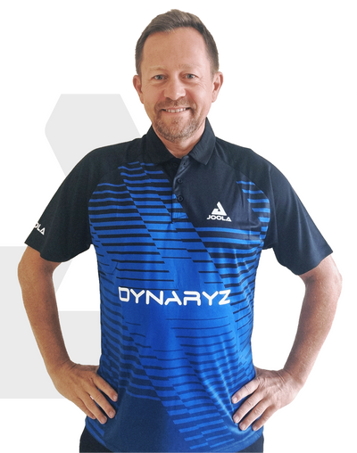 Mann trägt blau-schwarzes JOOLA Dynaryz Polo-Shirt mit kurzem Arm und Kragen, Hände in Hüfte, lächelnd.