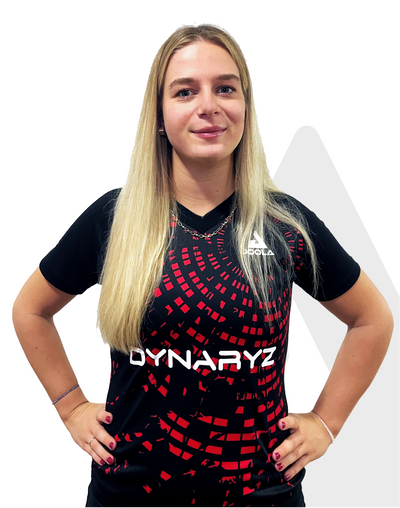 Frau mit langen blonden Haaren trägt schwarzes Sportshirt mit rotem Muster und weißem Dynaryz-Logo, Hände in Hüfte.