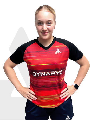 Frau trägt rotes und schwarzes JOOLA Dynaryz Sportshirt mit V-Ausschnitt und schwarzen Shorts, Hände in der Hüfte.