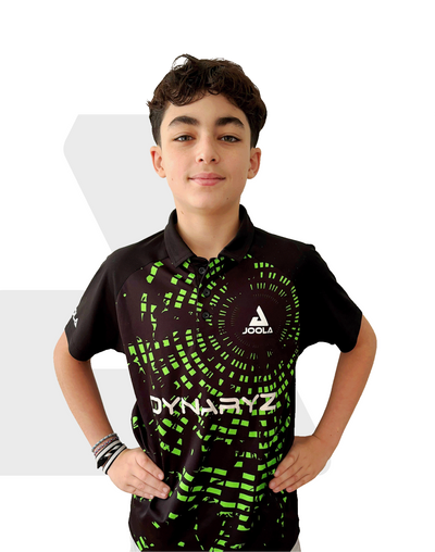 Junge trägt schwarzes JOOLA Dynaryz Poloshirt mit grünem Pixelmuster, Hände in der Hüfte, vor weißem Hintergrund.