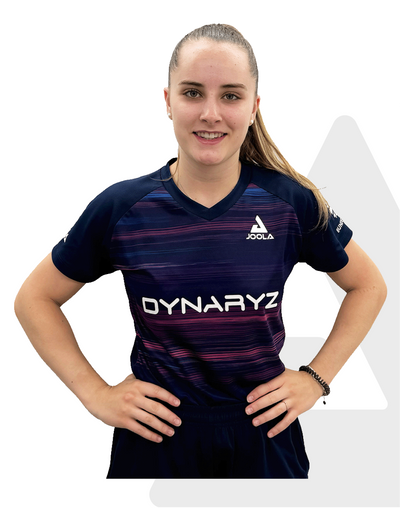Junge Frau mit Zopf trägt blau-lila Joola Dynaryz Sportshirt, Hände in der Hüfte, lächelnd vor weißem Hintergrund.