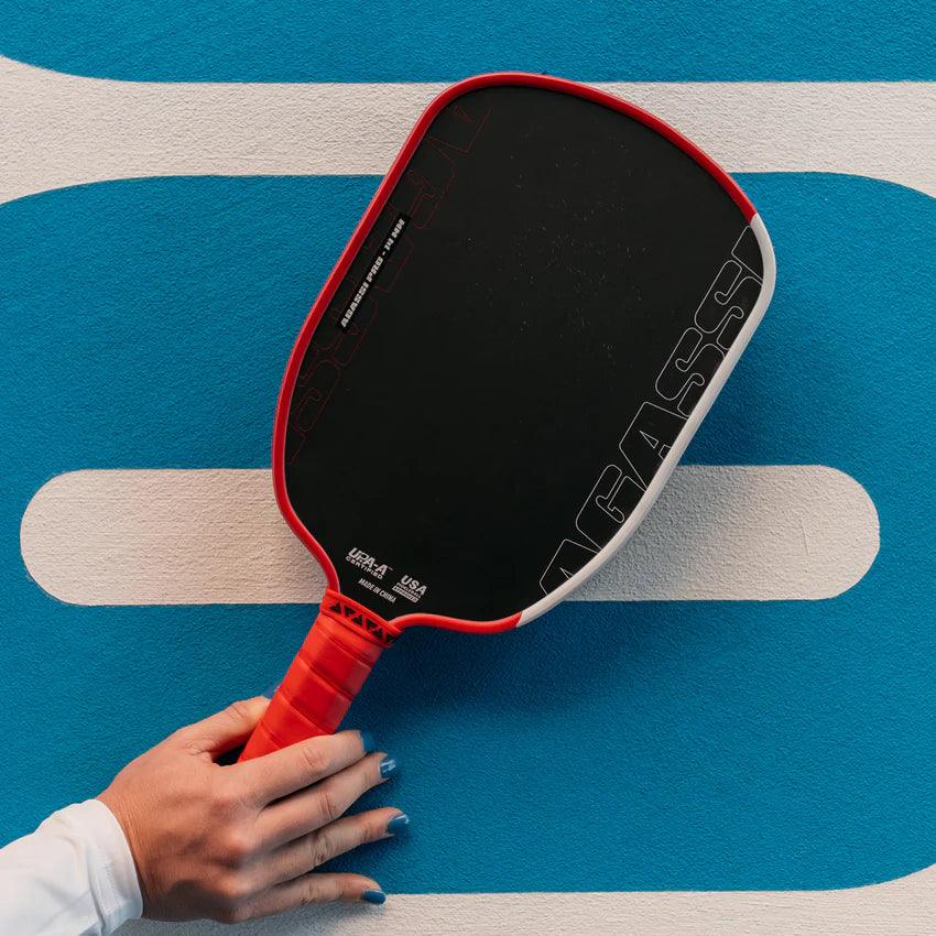 Hand mit rotem Griff hält schwarzen Pickleball-Schläger mit rotem Rahmen vor blau-weißem Hintergrund.