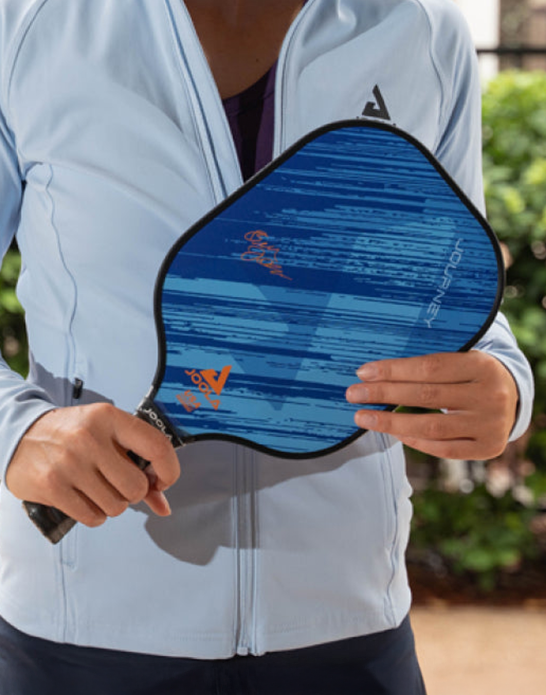 Person hält blauen JOOLA Pickleball-Schläger mit schwarzem Griff vor hellblauer Jacke, Outdoor-Hintergrund.