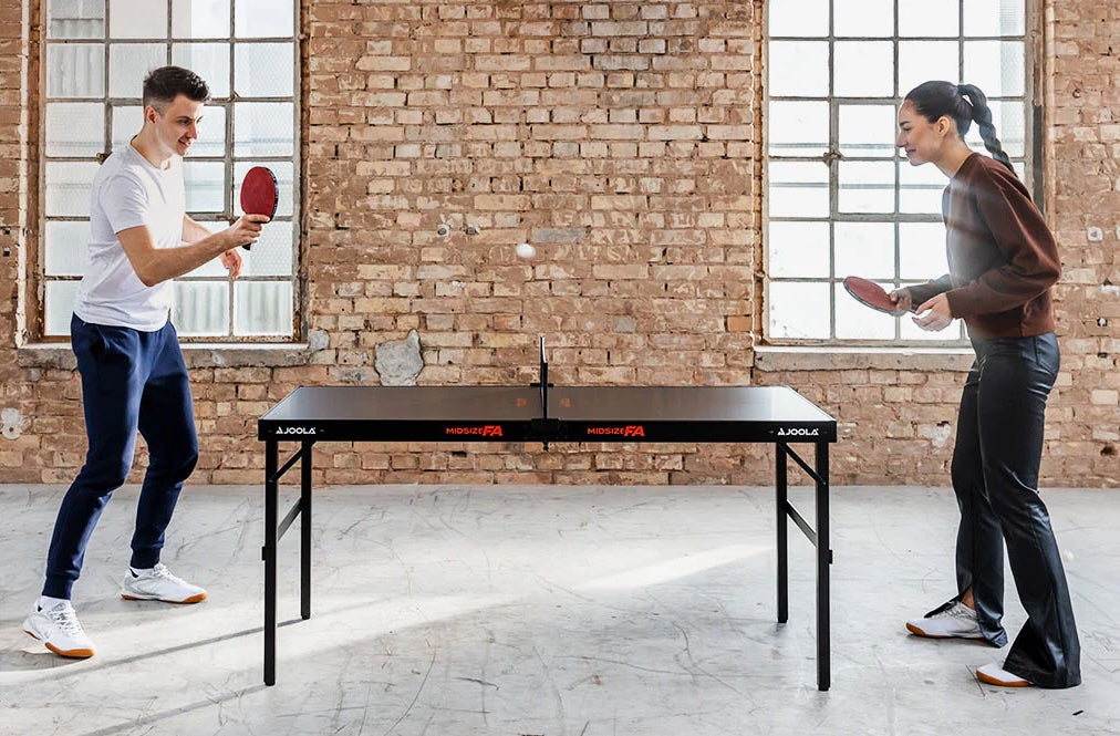 Zwei Personen spielen Tischtennis an einem schwarzen JOOLA Midsize FA Tisch in einem Raum mit Backsteinwand.