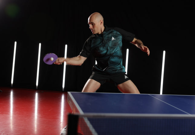 Mann in Sportbekleidung spielt Tischtennis, schlägt mit lila Schläger auf blauem Tisch vor dunklem Hintergrund.