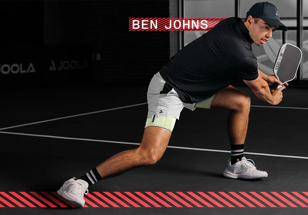 Mann in Sportkleidung mit Cap macht Ausfallschritt und hält Pickleball-Schläger auf schwarzem Spielfeld.