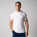Mann trägt weißes, kurzärmeliges Poloshirt mit kleinem schwarzem Logo auf der Brust und blauer Hose vor neutralem Hintergrund.