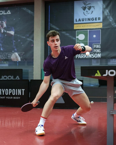 Junger Mann spielt Tischtennis, trägt lila Shirt und graue Shorts, dynamische Spielhaltung, roter Hallenboden.