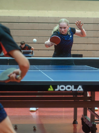 Frau spielt Tischtennis mit JOOLA Tischtennistisch, Schläger und Ball in Sporthalle mit Holzwand.
