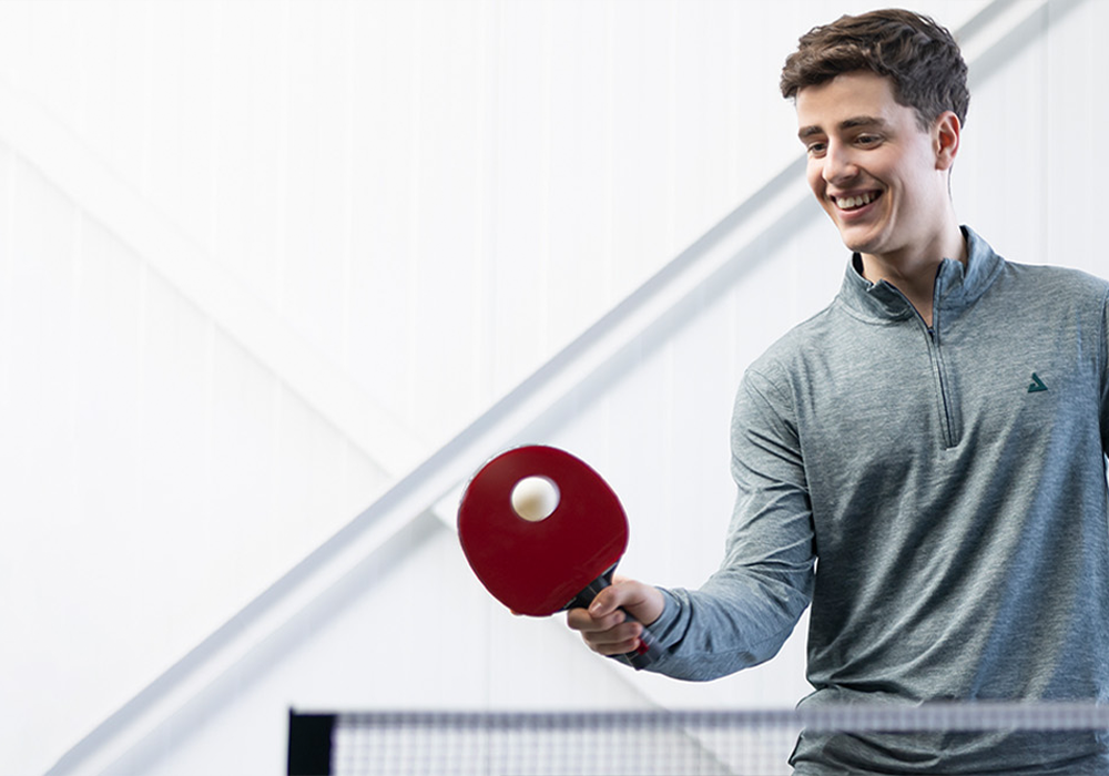 Junger Mann spielt Tischtennis mit rotem Schläger und weißem Ball vor hellem Hintergrund.