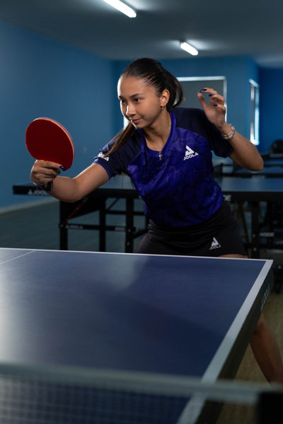 Frau in blauem JOOLA-Trikot spielt Tischtennis und hält roten Schläger vor blauem Tisch in Sporthalle.
