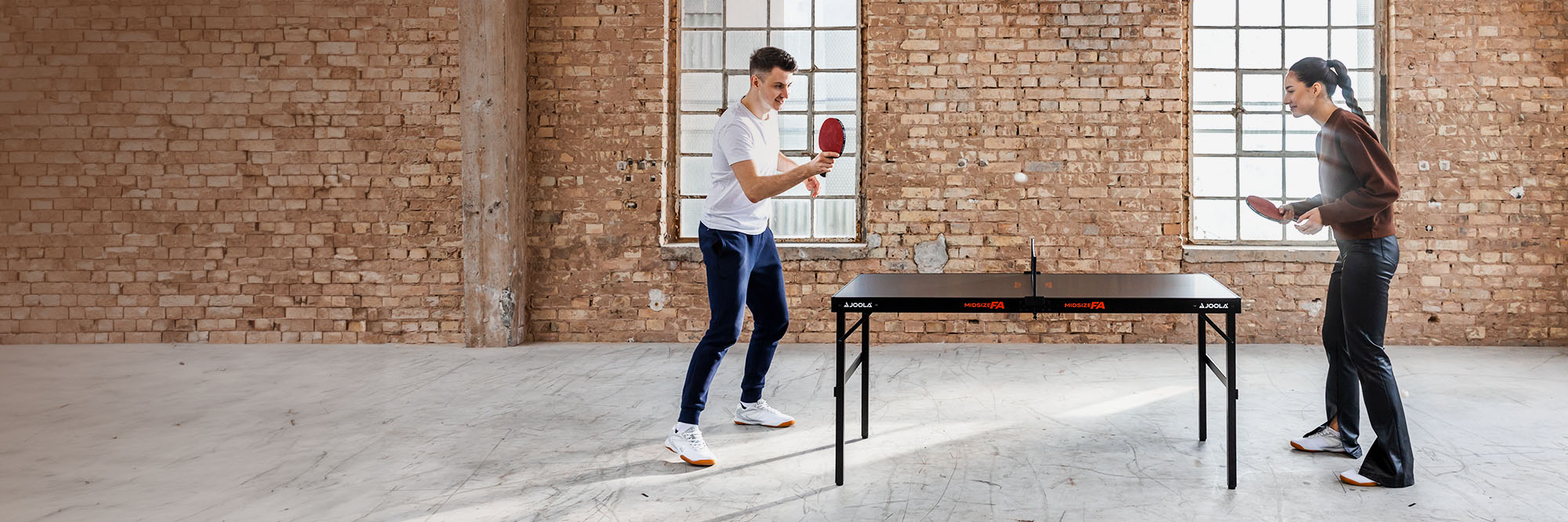 Zwei Personen spielen Tischtennis an einem schwarzen JOOLA Midsize FA Tisch in einem Raum mit Backsteinwänden.