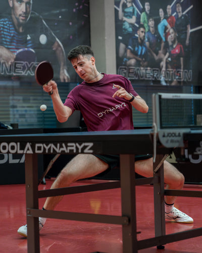 Mann in Sportkleidung spielt Tischtennis und schlägt Ball mit Schläger auf rotem Hallenboden.