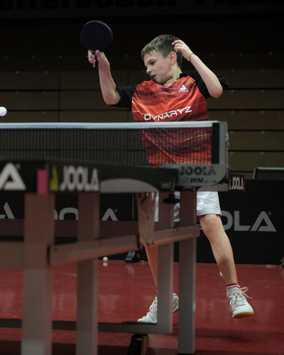 Junge in rotem Trikot spielt Tischtennis, schlägt Ball mit Schläger über Tisch mit Netz.